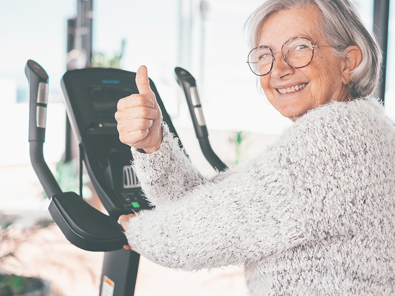 Happy senior woman doing exercises to keep fit by running on a stationary bike on the terrace of her house.